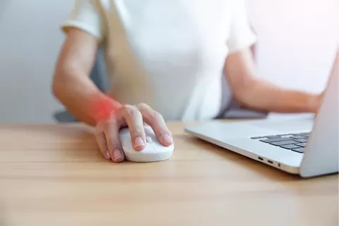 Afbeelding van een vrouw die voor haar computer zit en haar computermuis beweegt. Haar pols heeft een rode schijn om een pijn aan te duiden